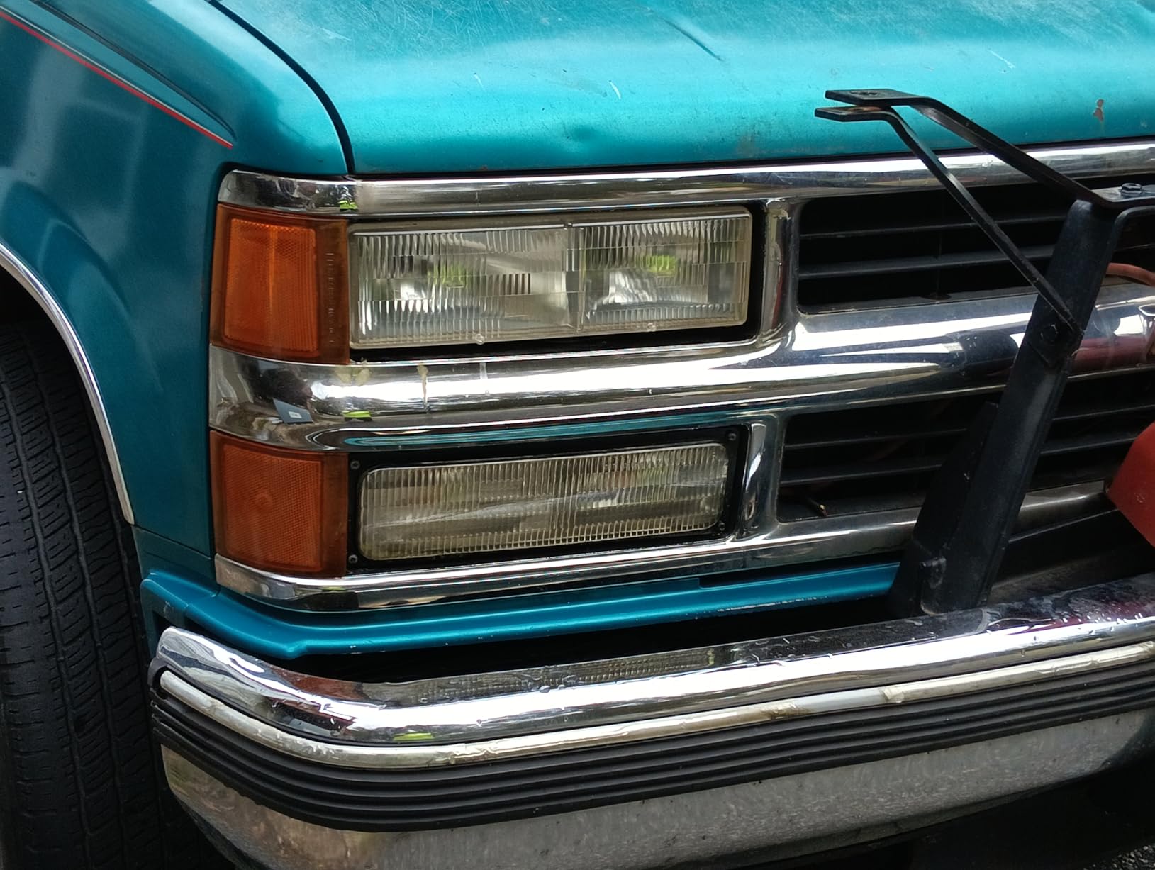 Front corner of a teal 1990s Chevrolet pickup with the upper headlight lens clear and the lower one still clouded